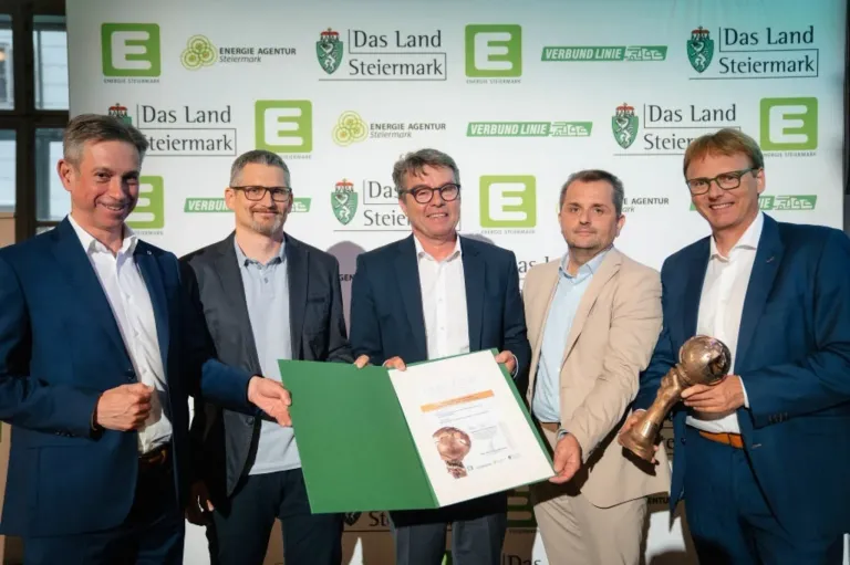 Fünf Männer stehen vor einer Wand mit Logos. Der Mann in der Mitte hält ein Zertifikat und Papier. Der Mann rechts hält eine Trophäe.