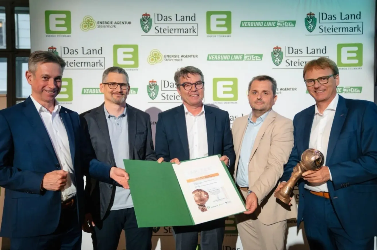 Fünf Männer stehen vor einer Wand mit Logos. Der Mann in der Mitte hält ein Zertifikat und Papier. Der Mann rechts hält eine Trophäe.