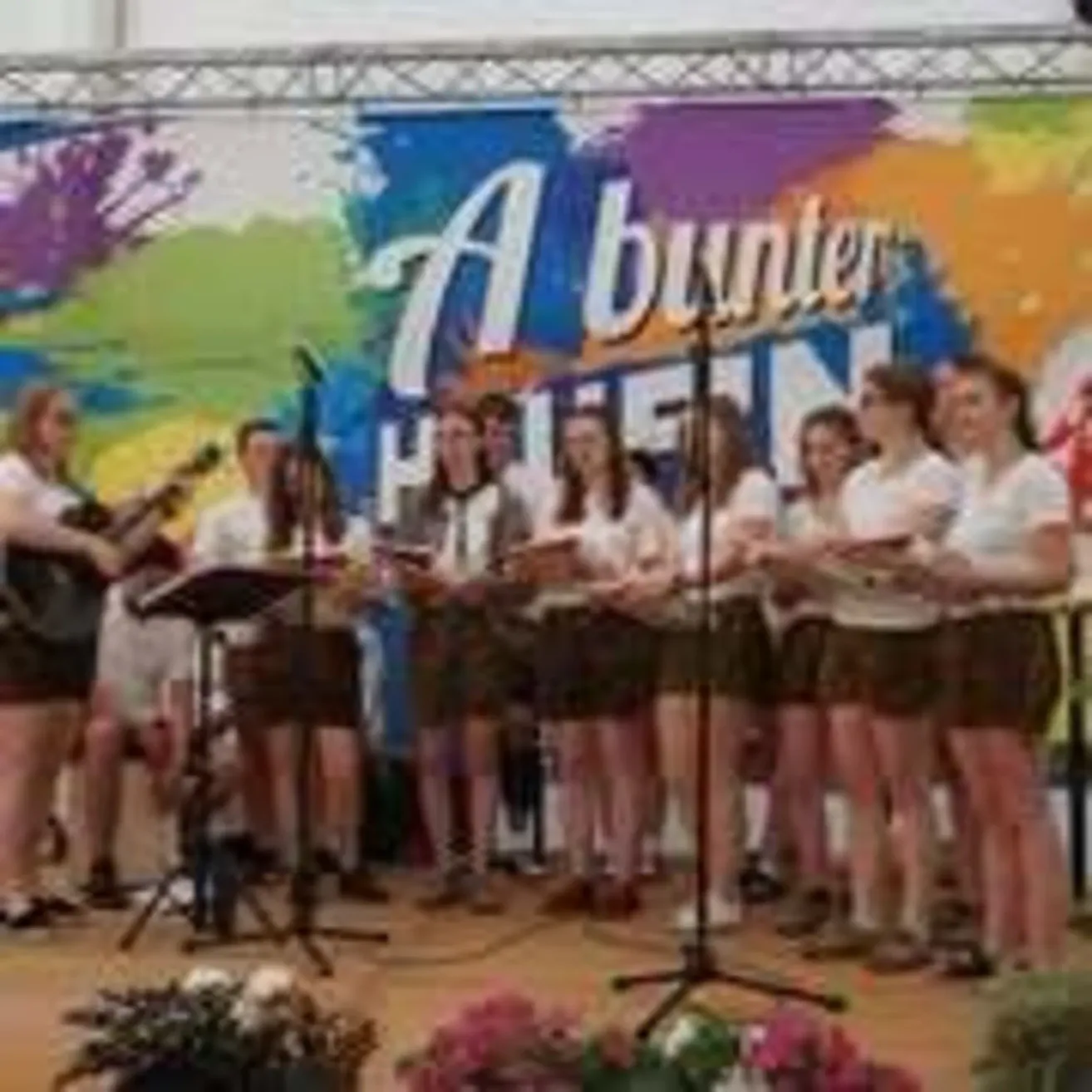 A choir on stage performs with musical instruments; a woman plays the guitar. A colorful backdrop features the word 'Abunter'.