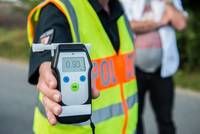 A person in a reflective vest holds a breathalyzer, likely conducting a sobriety test on a roadside.