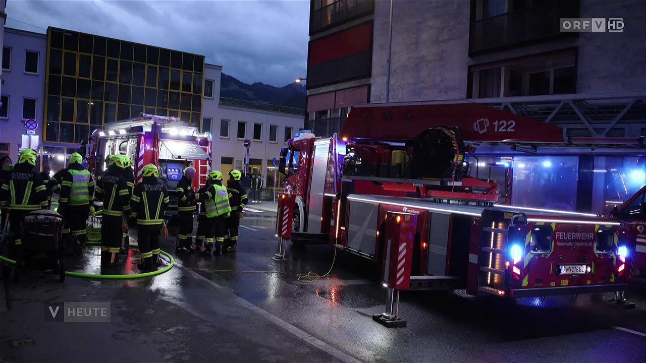 Firefighters are gathered around a fire truck on a wet road. The trucks have their lights on, and one has the number 122. Nearby, there are buildings and mountains.
