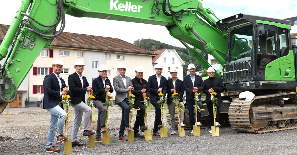 A group of men in suits and helmets hold shovels for a ground-breaking ceremony in front of a green Keller excavator.