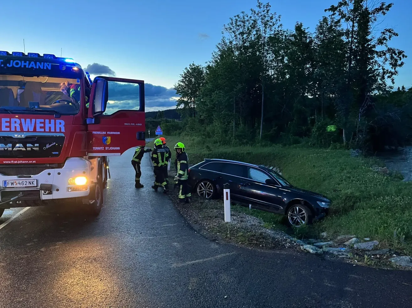 A fire truck is parked on the road. Three firefighters are standing around a damaged car on the roadside. The car is positioned on a grassy area.