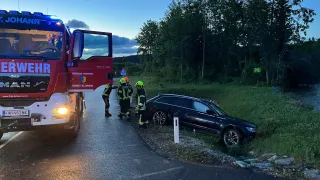 A fire truck is parked on the road. Three firefighters are standing around a damaged car on the roadside. The car is positioned on a grassy area.