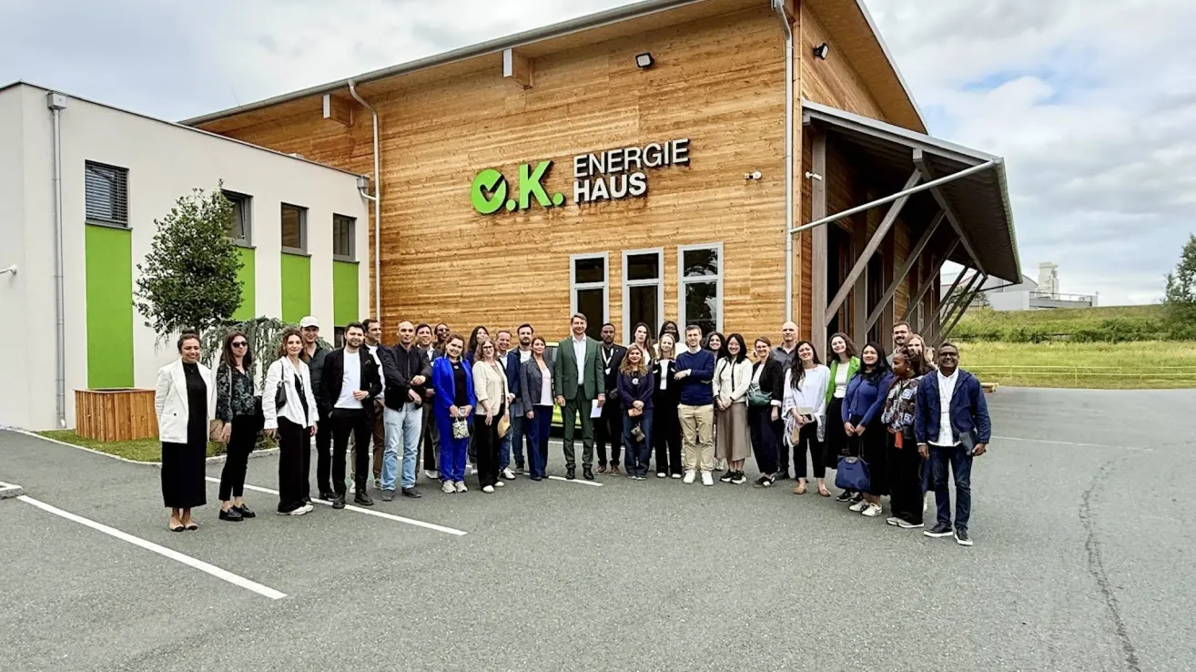 Eine Gruppe von Menschen steht vor einem Gebäude. Das Gebäude hat ein Logo und den Namen O.K. Energie Haus. Die Menschen sind in formeller Kleidung. Einige halten Taschen. Der Parkplatz ist leer.