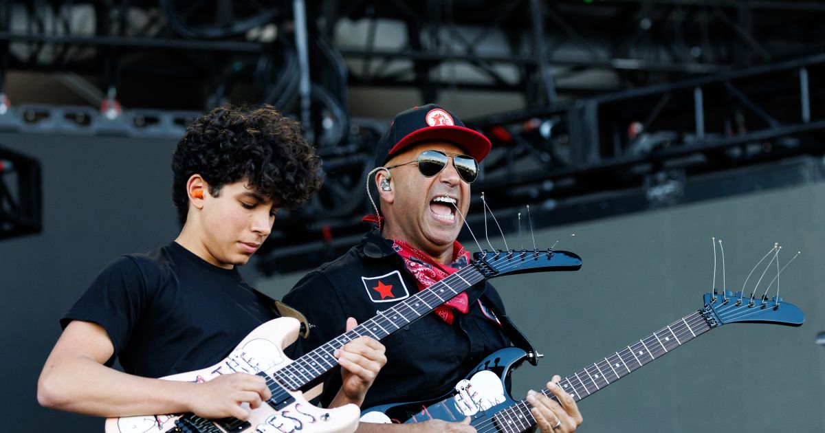Ein Mann in einem schwarzen Shirt mit rotem Halstuch und Sonnenbrille spielt Gitarre mit einem jungen Jungen. Der Mann singt in ein Mikrofon.