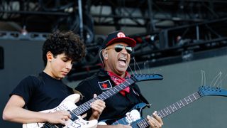 A man in a black shirt with a red bandana and sunglasses plays guitar with a young boy. The man is singing into a microphone.
