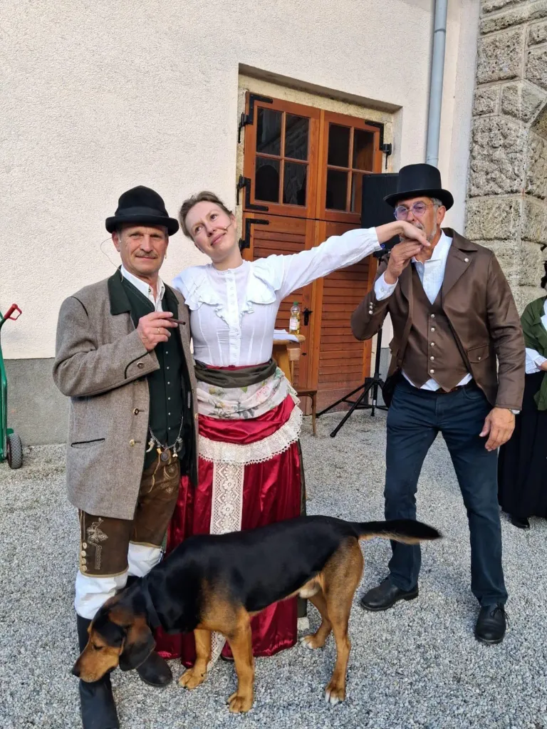 Three people in traditional dress, two men and a woman, pose with a black and brown dog in front of a building. The woman holds a cigar.
