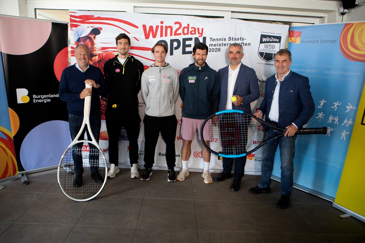 Five men stand in front of a banner, holding tennis rackets. They are wearing jackets and sneakers. One man holds a yellow tennis ball.