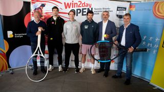 Five men stand in front of a banner, holding tennis rackets. They are wearing jackets and sneakers. One man holds a yellow tennis ball.