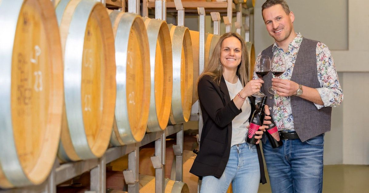 A man and a woman smile as they hold wine bottles in a wine cellar with large wooden barrels.