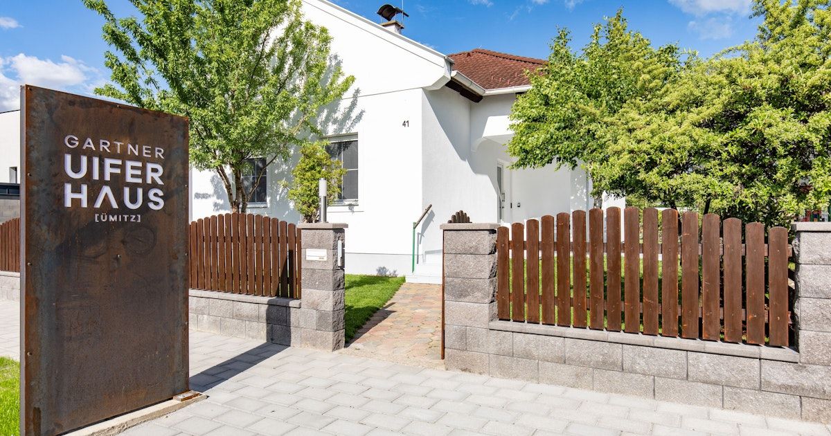 A modern, white house with a brown roof. A pathway leads to the front door, flanked by stone pillars and a wooden fence. Trees and greenery surround the house.