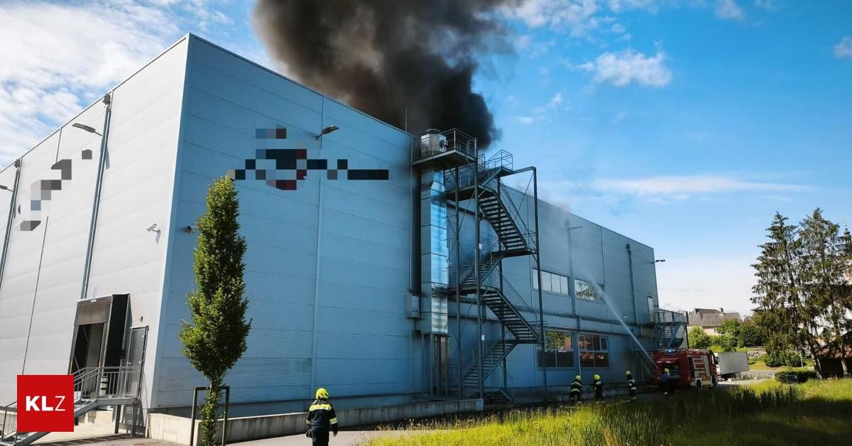 Firefighters extinguish a fire at a warehouse, with thick smoke billowing from the roof. A fire truck is parked nearby.