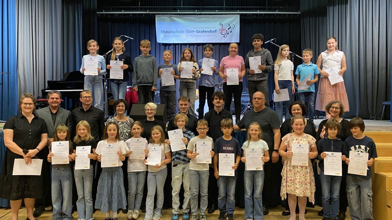 A group of students and teachers on stage, holding certificates, some wearing glasses. The backdrop includes a banner with the name 'Musikschule Ober-Grafendorf'. Microphones and speakers are set up.