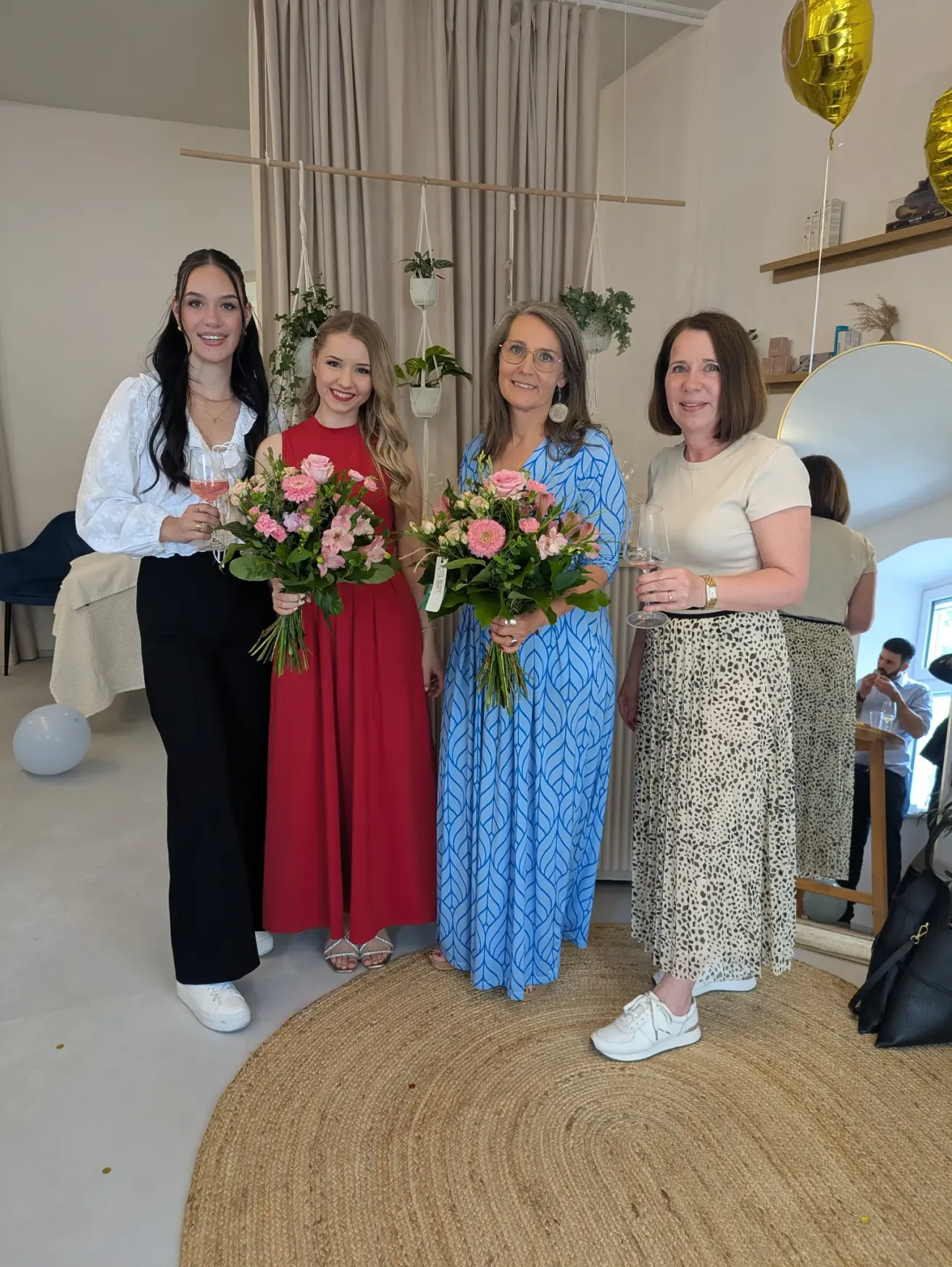 Vier Frauen stehen zusammen und halten Blumensträuße und Weingläser. Sie lächeln, und eine trägt eine Brille. Hinter ihnen ist ein Spiegel, in dem sich ein Mann spiegelt. Ein Stuhl und ein weißer Ball befinden sich im Hintergrund.