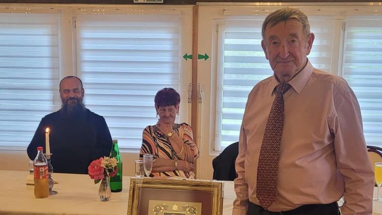 Ein älteres Paar steht neben einer gerahmten Auszeichnung. Eine Frau mit einer Blume in einer Vase und eine Flasche auf dem Tisch neben ihr. Dahinter sind Fenster mit Jalousien.