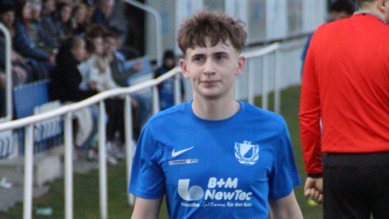 A soccer player in a blue shirt with a white Nike logo is standing on the field. In the background, there are blurry spectators.