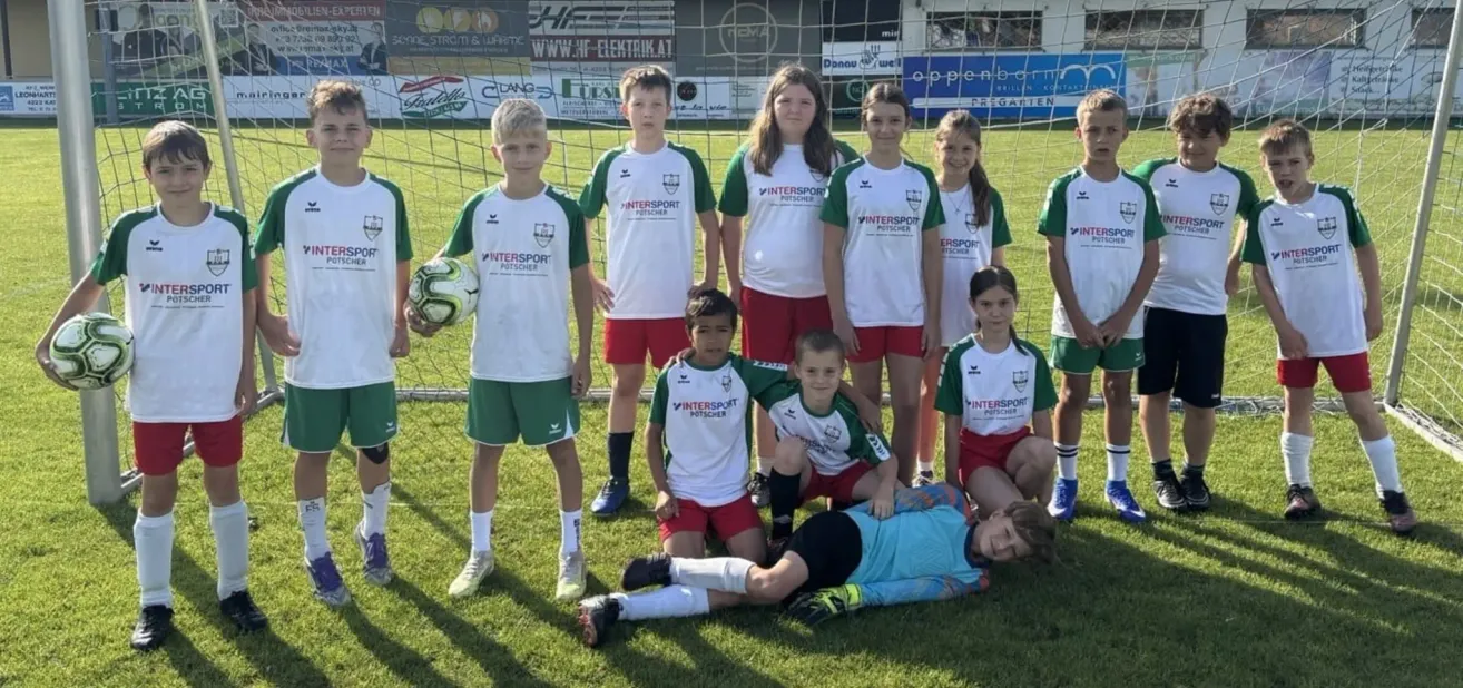 Eine Gruppe junger Fußballspieler posiert für ein Foto auf einem Feld mit einem Fußballnetz im Hintergrund. Sie tragen weiße und grüne Uniformen mit Intersport-Logo.