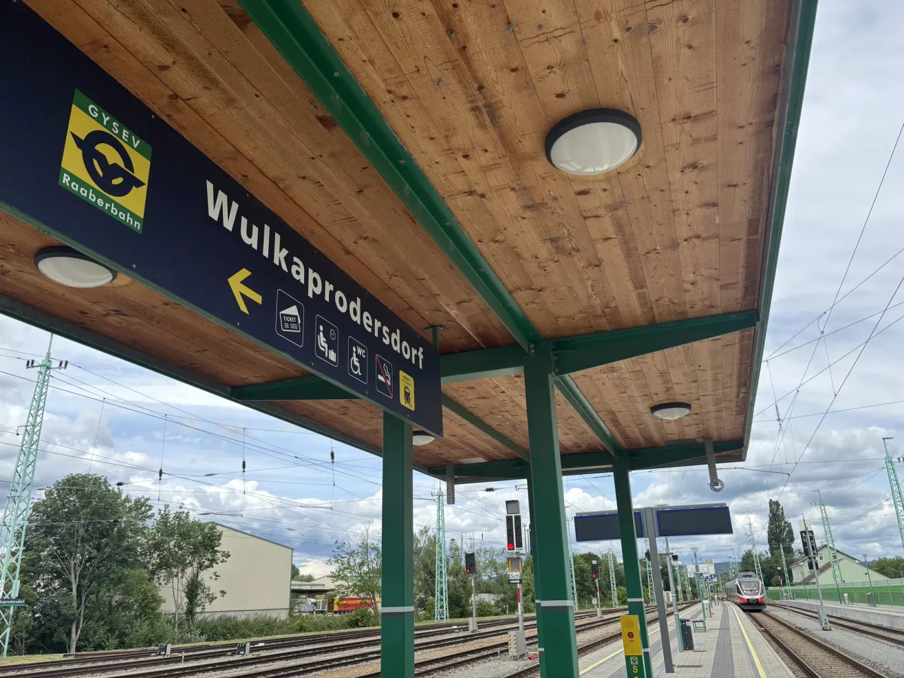 Eine Bahnsteigplattform mit einem Schild, das auf Wulkaprodersdorf hinweist. Der Bahnsteig hat eine Holzdecke, grüne Säulen und Deckenbeleuchtung. Gleise verlaufen parallel zum Bahnsteig.