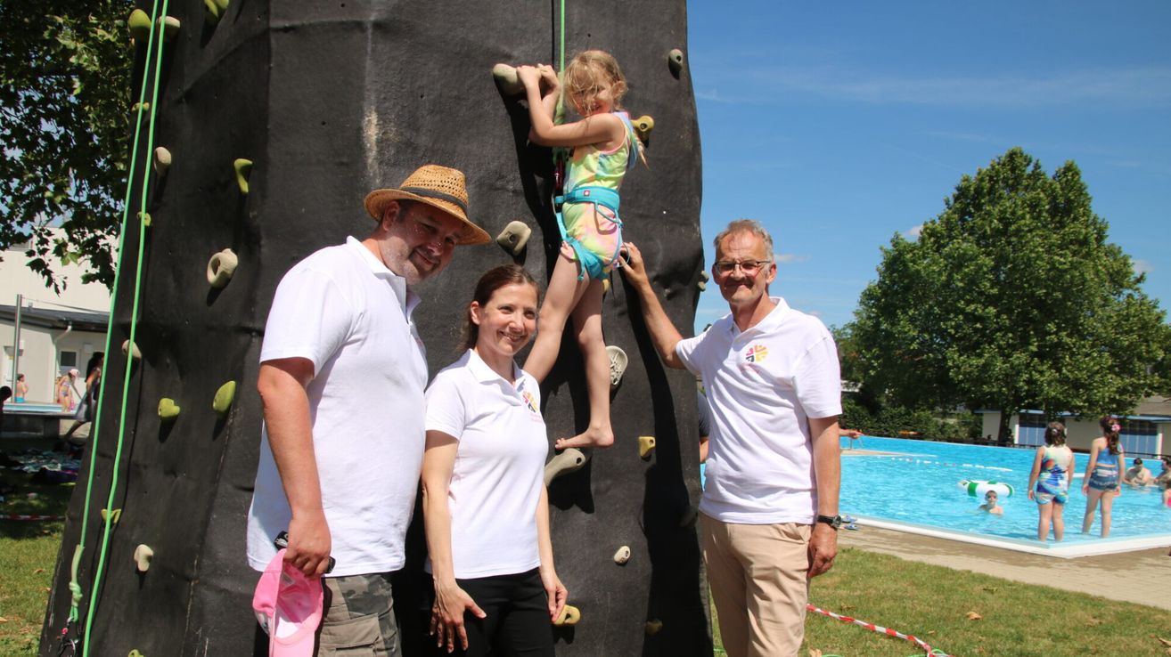 A young girl in a harness climbs a climbing wall with the support of two adults, one holding her from behind and the other holding her hand. They are all smiling and standing in front of a pool.