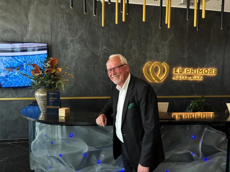 A smiling man in a suit stands in front of a hotel reception. Behind him is a wall with a logo, a flower arrangement, and a TV.
