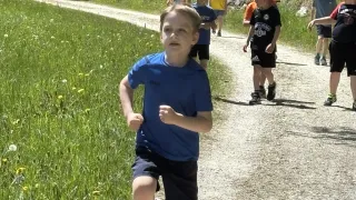 A group of children run along a dirt path with green grass on either side. They are wearing casual clothes and sneakers.