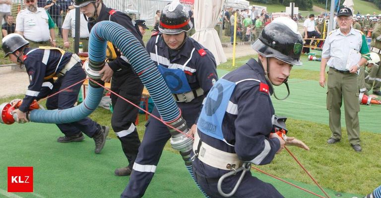 Drei Feuerwehrleute sind an einer Trainingsübung auf einem grünen Feld beteiligt. Sie benutzen einen großen Schlauch, und einer von ihnen hält ein Werkzeug. Die anderen beobachten.