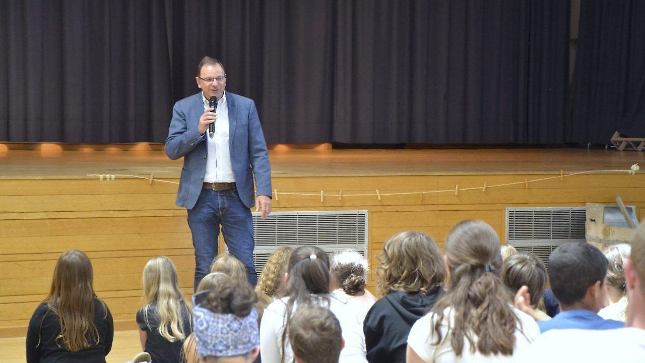 Ein Mann auf der Bühne mit einem Mikrofon spricht eine Gruppe von Schülern an. Er trägt einen blauen Anzug und eine Brille. Die Schüler sitzen vor ihm, einige tragen Stirnbänder.