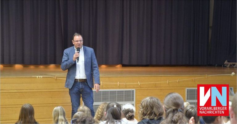 Ein Mann in einem blauen Anzug steht auf der Bühne und spricht eine Gruppe von Kindern mit einem Mikrofon an. Die Bühne hat schwarze Vorhänge im Hintergrund.