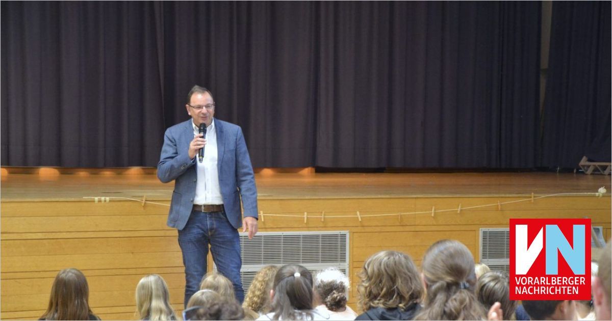 Ein Mann in einem blauen Anzug steht auf der Bühne und spricht eine Gruppe von Kindern mit einem Mikrofon an. Die Bühne hat schwarze Vorhänge im Hintergrund.