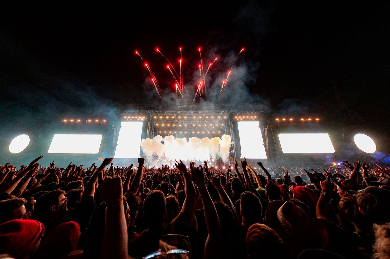 Ein großer Haufen versammelt sich unter dem Nachthimmel und sieht ein Konzert mit roten Feuerwerken, Rauch und hellen Bühnenlichtern. Das Publikum hebt die Hände und jubelt.