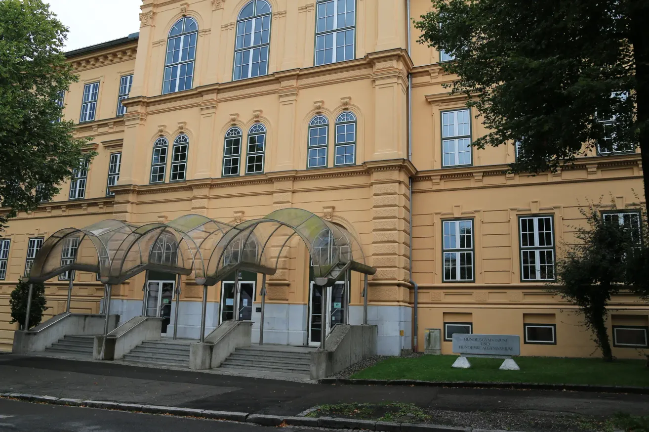 A large yellow building with many windows and glass arches over the entrance. It has a grassy lawn in front and a sign reading Bundesgymnasium.