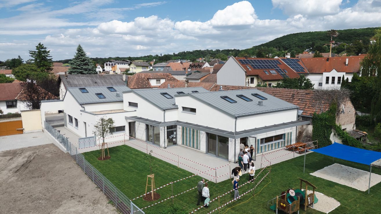 Aerial view of a modern building with solar panels on the roofs. People are walking around, some are entering the building. Trees and plants are in front of the building.