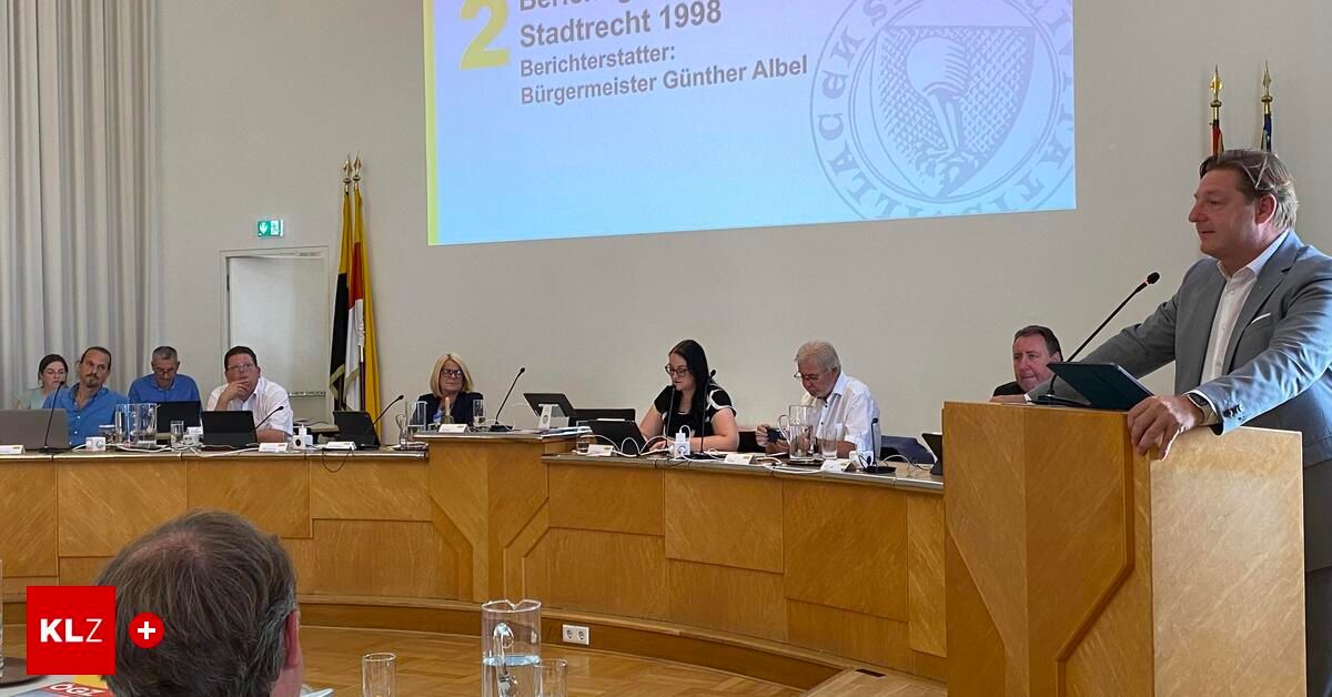Five people sit around a table in a meeting room, facing a projector screen displaying 'Stadtrecht 1998'. A woman with glasses sits next to a man wearing glasses. A flag is on the wall behind them.