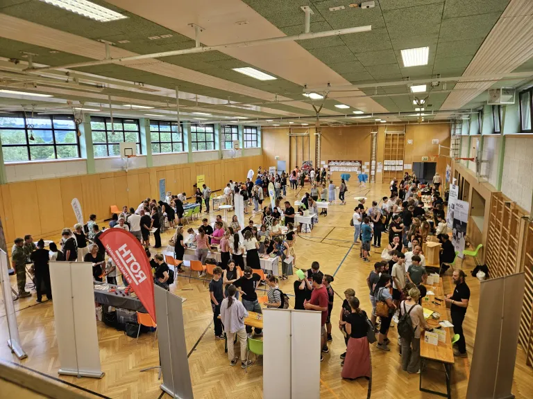 Ein großer Innenraum mit vielen Menschen auf einer Jobmesse. Die Menschen versammeln sich um Tische und Stände, führen Gespräche und stöbern in Materialien. Der Boden ist aus Holz und die Wände sind aus Holz.