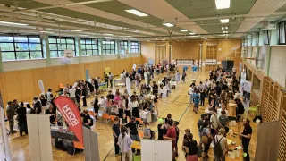 A large indoor space with many people at a job fair. People are gathered around tables and booths, engaging in conversations and browsing through materials. The floor is wooden, and the walls are made of wood.