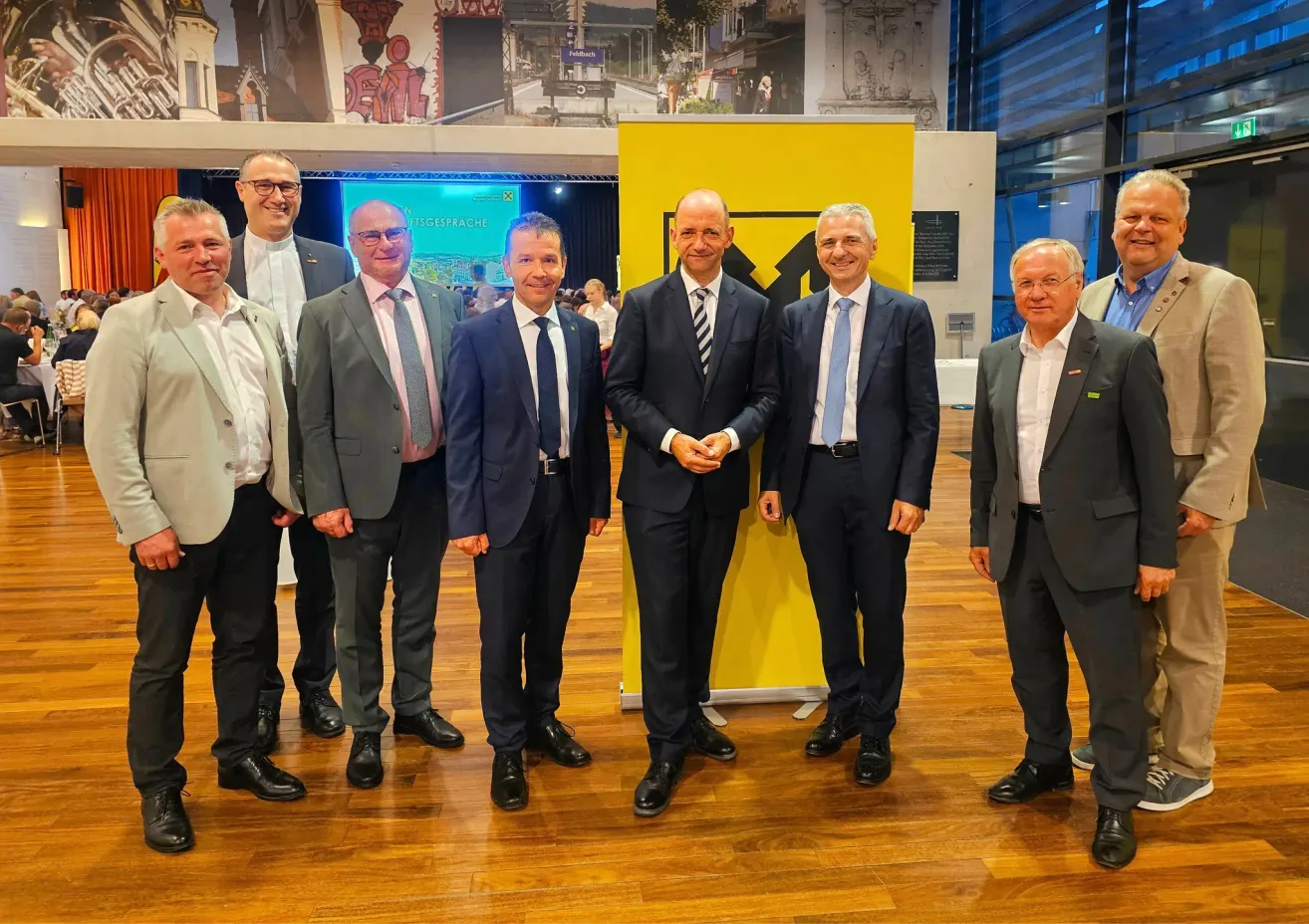 Seven men in suits stand in a row on a wooden floor. A yellow banner with black writing is behind them. They appear to be posing for a photograph.