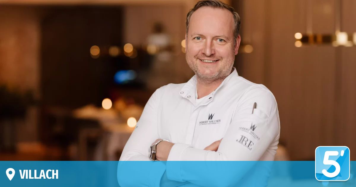 A smiling man in a chef's uniform with a name tag and a watch, standing with his arms crossed in a restaurant.