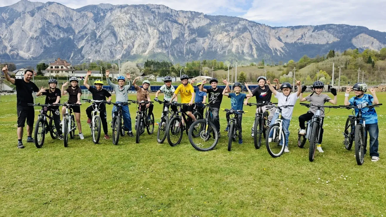 Eine Gruppe junger Jungen und Mädchen mit Helmen und Turnschuhen posiert für ein Foto auf einem Rasenfeld mit Fahrrädern vor einer Bergkette.