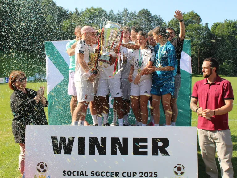 Fußballmannschaft feiert auf einem Podium mit Konfetti und einem Pokal in der Hand. Das Schild steht 'WINNER SOCIAL SOCCER CUP 2025'.