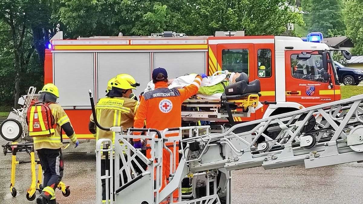 Emergency personnel in orange uniforms transfer a patient on a stretcher to an ambulance. A firefighter in a yellow helmet stands behind them. Trees and a red fire truck are in the background.