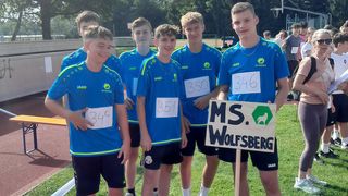 A group of young athletes wearing blue shirts with numbers on them are standing on a track. They are smiling and posing for a photo with a sign reading 'MS. Wolfsberg'. Behind them, more people are standing on the grass.
