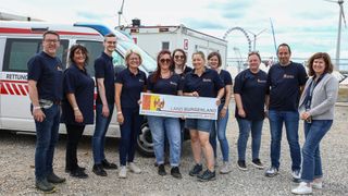 A group of smiling people in matching uniforms pose in front of a van and a large banner reading Land Burgenland. Behind them is a Ferris wheel and a cloudy sky.