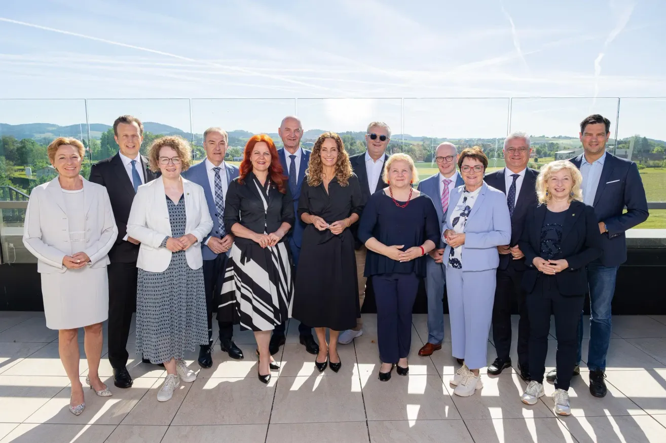 Eine Gruppe von Menschen steht zusammen, möglicherweise für ein Foto. Sie sind in formeller Kleidung gekleidet. In der Ferne sind Berge zu sehen, und der Himmel ist klar.