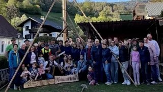 A large group of people poses for a photo in front of a wooden structure. Some hold wooden sticks, and some wear hats. Two wooden signs with the text 'Kuhstall' are on the ground. A house with a chimney is behind them.