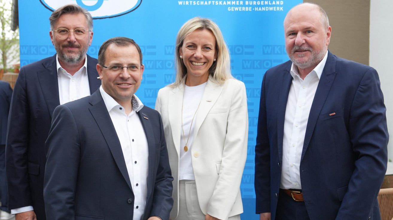 Three people dressed in formal attire stand before a blue backdrop. A woman smiles in the middle, and two men stand on either side of her. Behind them, there is a logo in white and blue.