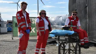 Three people in red safety suits stand outside near a wheeled cart with a blue cover and a backdrop of windmills.