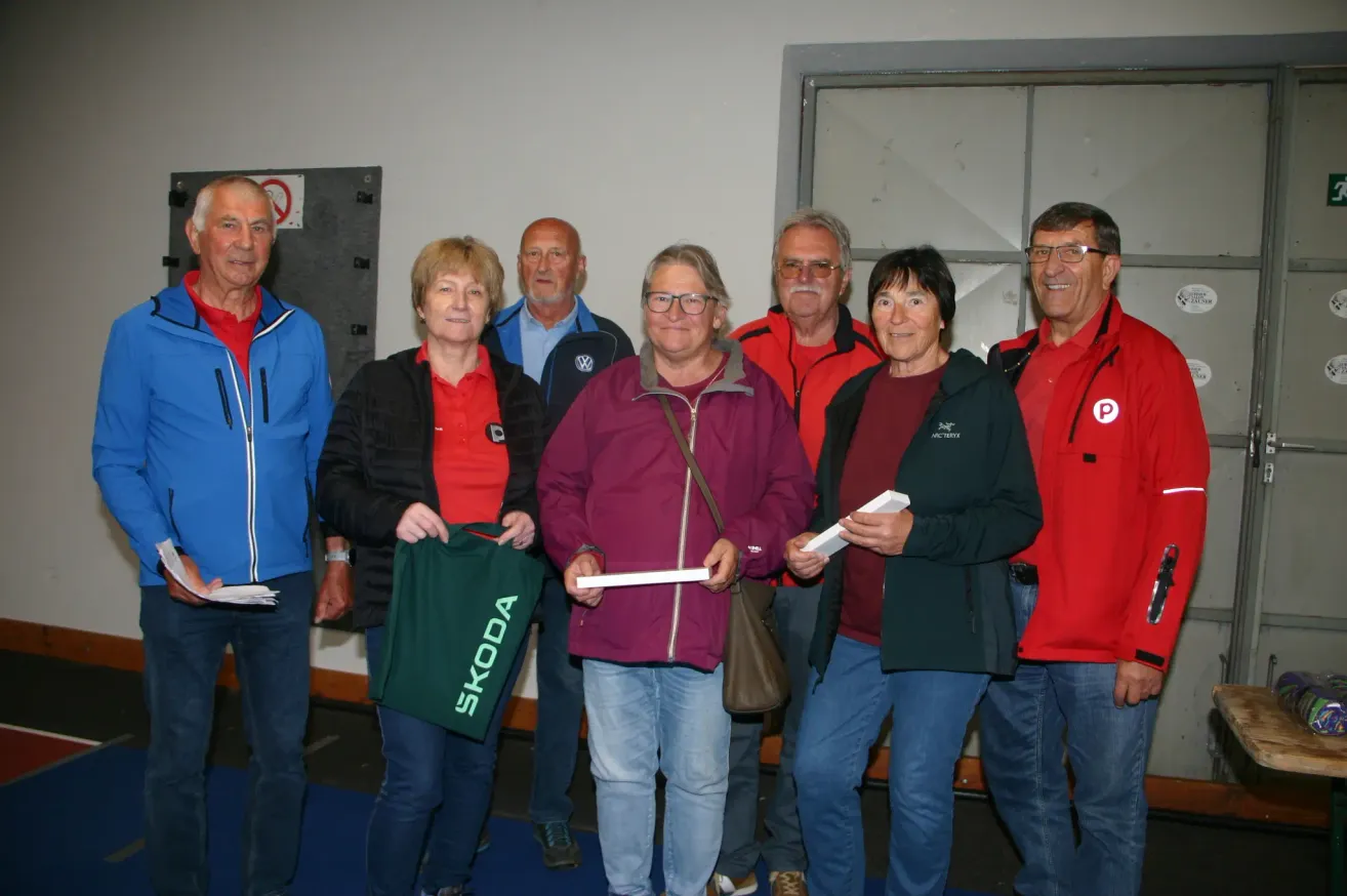 Eine Gruppe von Menschen in Jacken posiert für ein Foto. Eine Frau hält einen grünen Stoff mit dem Wort Skoda. Eine andere Frau hält ein weißes Papier. Sie alle tragen eine Brille. Hinter ihnen ist eine weiße Wand mit einem Glasfenster.