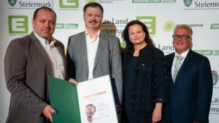 Three individuals stand together holding an award certificate. The man on the left holds a folder with the certificate inside. They stand in front of a wall with logos and text.