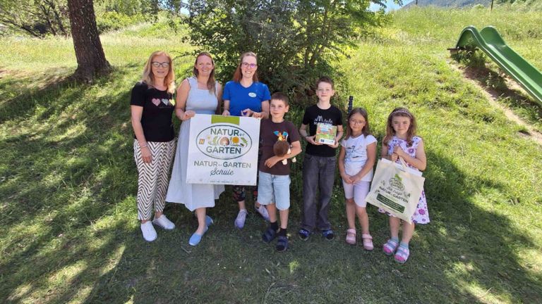 Eine Gruppe von Erwachsenen und Kindern steht auf einem Feld, lächelt und hält ein weißes Banner mit der Aufschrift 'Natur-Garten'. Sie tragen Taschen und scheinen für ein Foto zu posieren.
